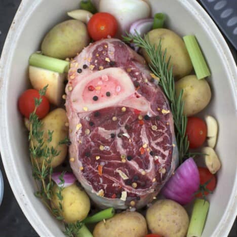 A raw beef shank sits in a white casserole dish, surrounded by uncooked potatoes, onions, tomatoes, and fresh herbs.