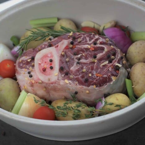 A raw beef shank is seasoned with whole peppercorns, fresh herb sprigs, and is nestled among yellow potatoes, carrots, and onions, in a white casserole dish.