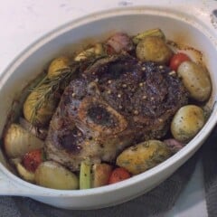 A roasted beef shank sits in a white oval casserole dish, surrounded by cooked potatoes, carrots, and cherry tomatoes.