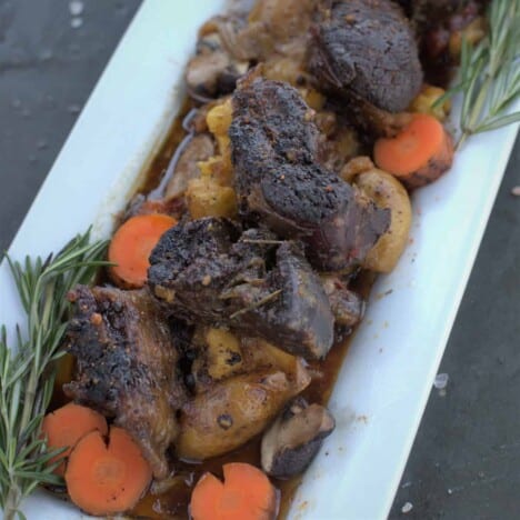 Cooked and sliced beef shank is layered on top of roasted potatoes, onions, and carrots in a white rectangular serving platter.
