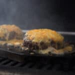 Two hamburgers sit on a grill, smothered in melting shredded cheddar cheese.