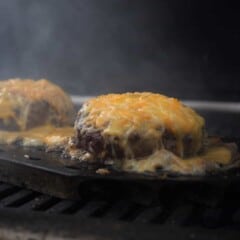 Two hamburgers sit on a grill, smothered in melting shredded cheddar cheese.