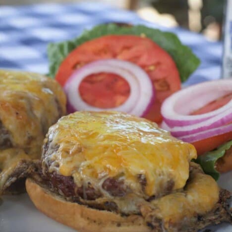 A cheeseburger on a bun, with sliced red onions and tomatoes, sits atop a blue and white checkered tablecloth.
