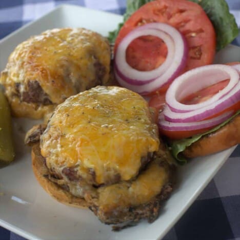 Looking down onto two cheeseburgers on a white plate, served with red onion and tomato slices.