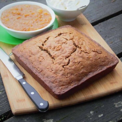 A large square carrot cake sits on a wooden cutting board with a green bowl of pineapple sauce and a knife by its side.