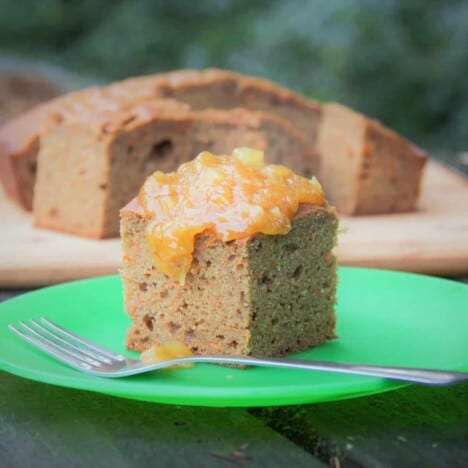 A square piece of carrot cake with a dollop of pineapple sauce sits on a round green plate with a fork, with additional squares of cake in the background.