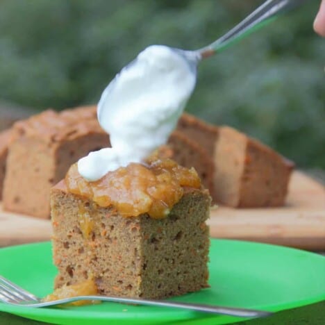 A spoon places a dollop of whipped cream on top of a square piece of carrot cake sitting on a bright green plate.