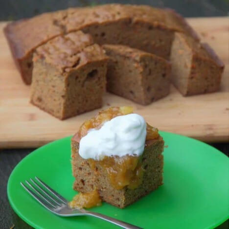 A square piece of carrot cake, dolloped with whipped cream, sits on a round green plate, with additional squares of carrot cake in the background.