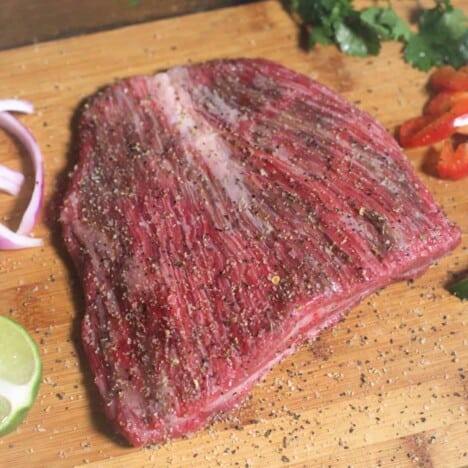 Looking down onto a flank steak sprinkled with seasoning, resting on a wooden cutting board.