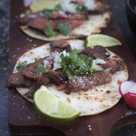 Looking down on an assembled taco with grilled flank steak slices, cilantro, crumbled cotija cheese, and lime wedges, on a dark wood background.