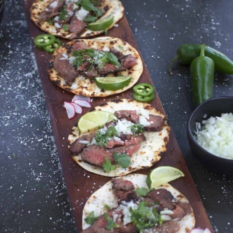 Looking down on a row of assembled tacos, with flank steak, cilantro, diced onions, and lime wedges, on a dark blue background.