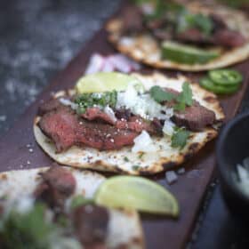 Looking down onto a charred tortilla sitting on a plank of wood, topped with grilled flank steak, cilantro, diced onions, and lime wedges.
