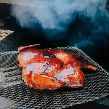 A glazed and butterflied chicken is cooking on a grill grate with smoke in the background.