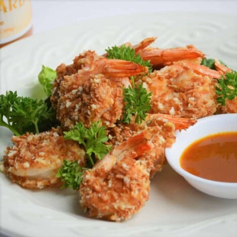 A white plate holds a pile of coconut shrimp garnished with parsley, served alongside a red dipping sauce.