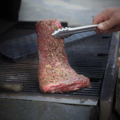 A raw flat iron steak is lowered onto a hot grill with a pair of metal tongs.