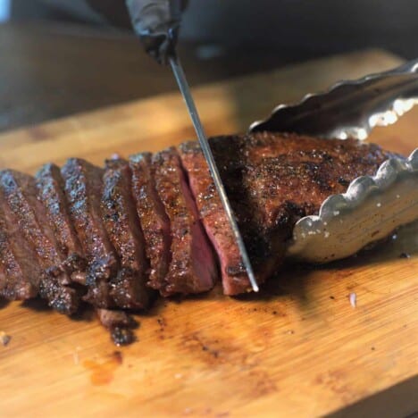 A grilled flat iron steak sits on a wood cutting board and is held by a pair of metal tongs while being thinly sliced against the grain.