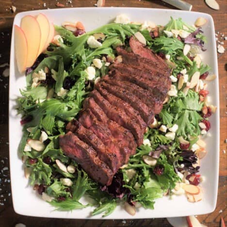 Looking down onto a large white square platter with sliced flat iron steak on top of a bed of lettuce sprinkled with feta cheese and dried fruit.