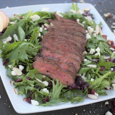 A grilled and sliced flat iron steak is layered on top of a bed of salad greens, sprinkled with feta cheese served on a square white serving platter.