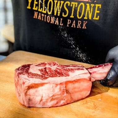 A raw tomahawk ribeye steak lays on a wooden cutting board, with a person in a "Yellowstone" black t-shirt behind it.