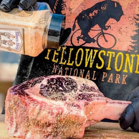 A person wearing a black t-shirt with the words "Yellowstone" on it is seasoning a raw tomahawk ribeye steak on a wooden cutting board.