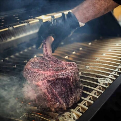 A person wearing a black glove is searing a raw tomahawk ribeye steak over a hot grill.