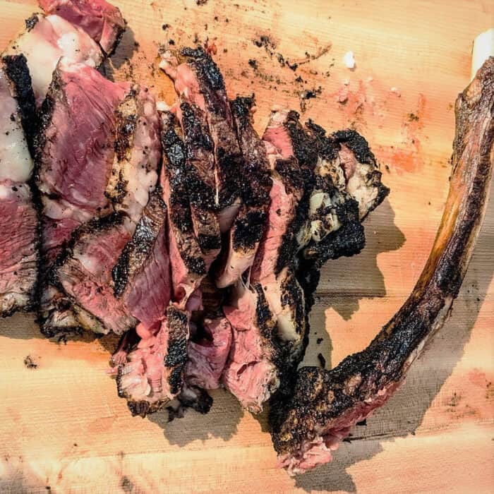 A grilled and sliced tomahawk ribeye steak is shingled on a wooden cutting board.
