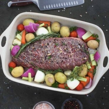 A seasoned London broil roast sits in a white roasting pan, surrounded by yellow potatoes, red onions, carrots, and other vegetables.