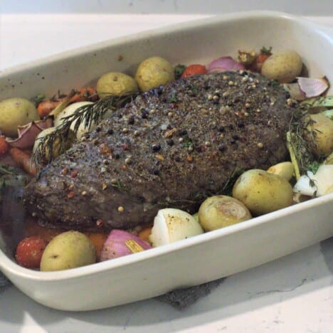 Looking into a rectangular white roasting pan holding a London broil roast surrounded by raw baby yellow potatoes and chopped red onions.