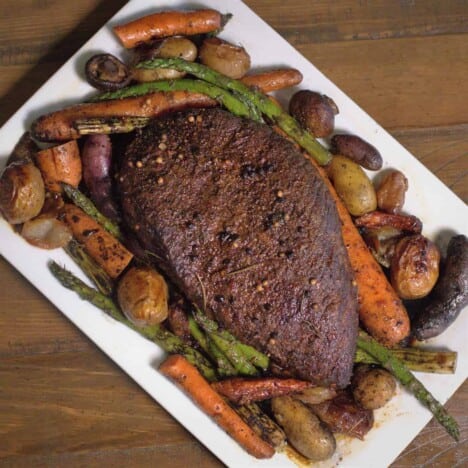 A slow cooked London broil roast sits on a rectangular white serving platter, with roasted potatoes, carrots, and fresh herbs.
