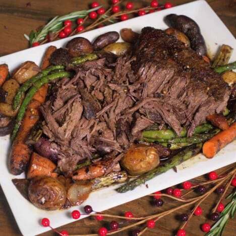 Shredded London broil roasts is piled on a rectangular white serving platter, surrounded by cooked yellow baby potatoes and carrots, with festive red berries and twigs on the table.