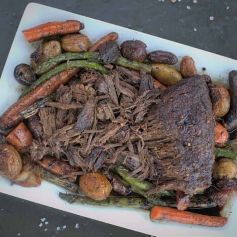 A cooked London broil roast sits on a rectangular white serving platter, surrounded by cooked potatoes, carrots, and green beans.