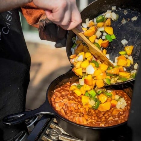 A cast iron skillet of chopped peaches, peppers, and onions are being transferred to a cast iron pot of baked beans on a grill.