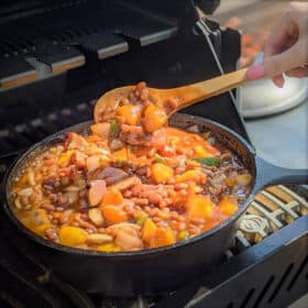 A cast iron pot of simmering baked beans, peaches, green bell peppers, and onions simmers on a grill, while being stirred by a wooden spoon.
