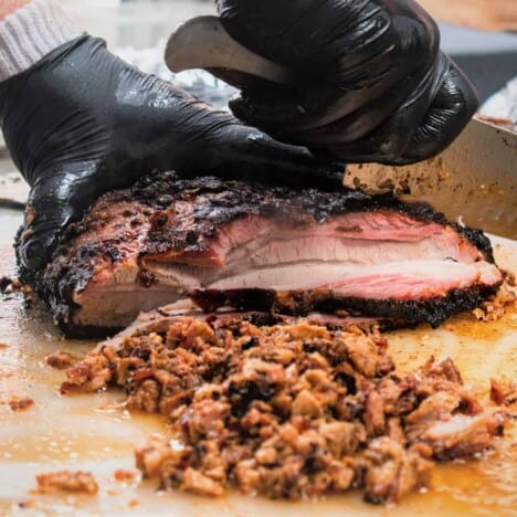 A slab of cooked pork belly with a dark brown crust is being diced into small pieces on a wooden cutting board.