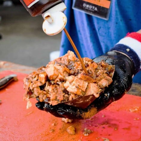 A hand wearing a black glove holds a pile of diced and sauced pork belly over a red cutting board.
