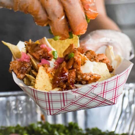 A white hand is reaching for a tortilla chip covered in diced pork belly, pickled onions, queso, and red sauce in a small red and white checkered paper plate.