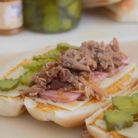 A close up of a pulled pork Cuban sandwich, open on a cutting board, with piles of pulled pork and pickles.