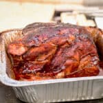 A smoked pork butt is slathered in barbecue sauce and is resting in a foil dish.