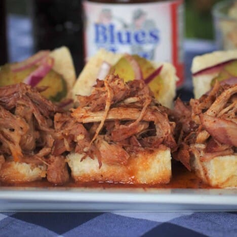 Open-faced slider buns are piled with pulled pork and topped with pickles, lined up in a rectangular white serving platter.
