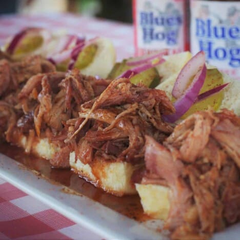 A row of shredded pork sliders topped with pickles and sliced red onions sit on a white serving platter with Blues Hog brand sauce and a red and white checkered tablecloth in the background.