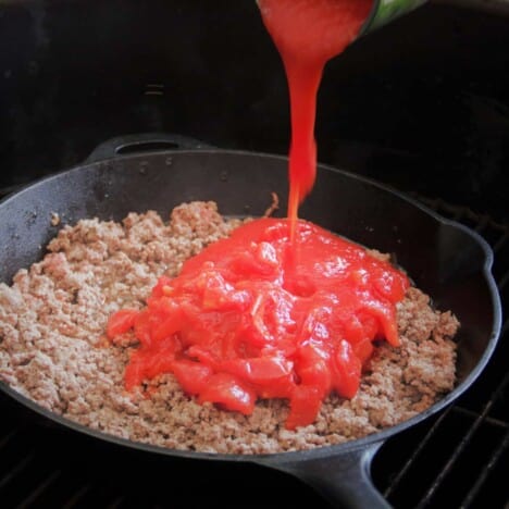 Tomato sauce is being poured over cooked brown been in a cast iron skillet on a grill grate.