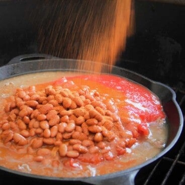 A cast iron skillet, sitting on a grill grate, is filled with ground beef, chili beans, and tomatoes, and is being sprinkled with seasoning.