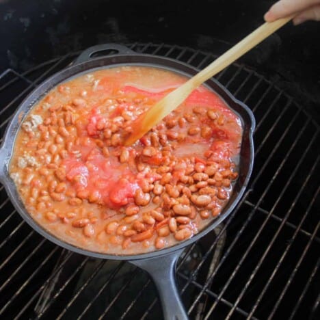 A cast iron skillet, sitting on a grill grate, is filled with a beef, bean, and tomato mixture being stirred by a wooden spoon.