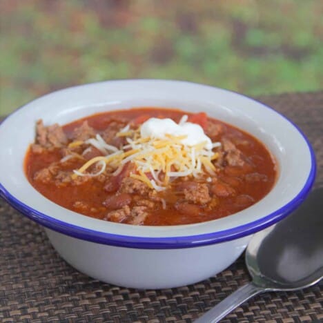 A white bowl with a blue rim is filled with beef chili with shredded cheese and sour cream on top.