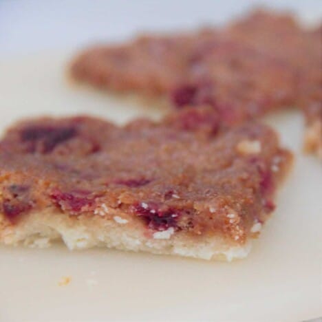 Looking across two pieces of raspberry slice, with a thin shortbread crust and a reddish-brown filling, on a cream-colored background.
