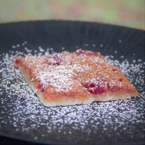 A piece of raspberry slice, with a thin shortbread crust, is dusted with confectioners sugar and is placed on a black plate.