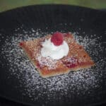 A large square of raspberry slice with a dollop of whipped cream, a fresh raspberry, and confectioners sugar sits on a black plate.