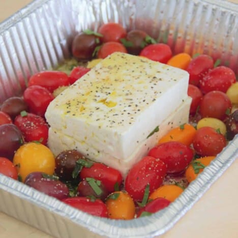 Looking into a rectangular foil cooking pan with two blocks of fresh, seasoned feta cheese surrounded by cherry tomatoes.