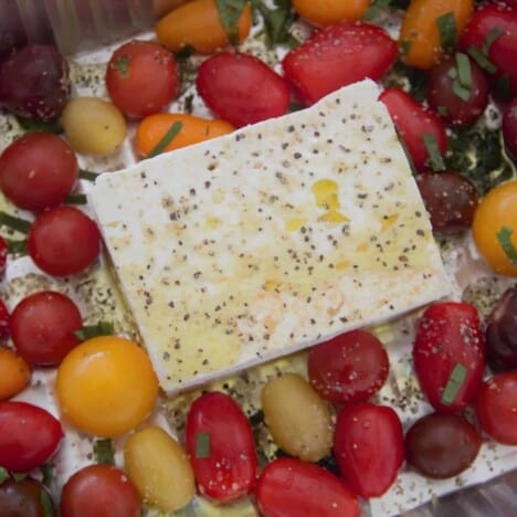 A close up shot of a block of feta cheese, drizzled with oil and sprinkled with pepper, nestled in cherry tomatoes.