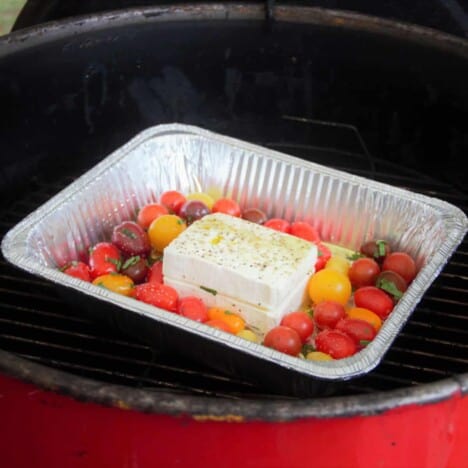 A block of feta cheese is nestled in cherry tomatoes in a foil baking dish on a grill.